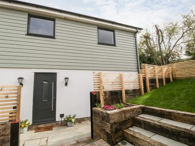 A house entrance with a black door and two windows above a stone patio with flower pots and wooden steps leading to a grassy slope at Sevenwoods View in Longhope