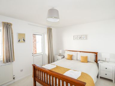 A bedroom with a wooden bed frame white bedding yellow cushions and runners two white bedside tables with lamps and a door with curtains at The Summerhouse in Y Felinheli