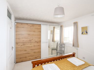 A bedroom with a wooden bed frame and mustard quilt a chair in front of a mirrored wardrobe and windows with beige curtains at The Summerhouse in Y Felinheli