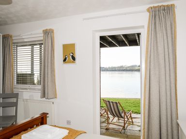 A bedroom with a bed and stacked towels near a window and open door showing deck chairs outside by water at The Summerhouse in Y Felinheli