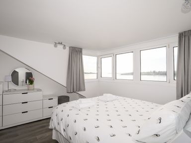 A bedroom with a bed, dresser with mirror, lamp, and large windows with gray curtains at The Summerhouse in Y Felinheli