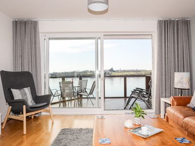 A living room with a wooden floor, gray armchair, brown leather sofa, coffee table, and sliding glass door leading to a balcony overlooking water at The Summerhouse in Y Felinheli