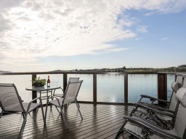 A wooden deck with chairs a round table with a bottle and glasses overlooking a body of water at The Summerhouse in Y Felinheli