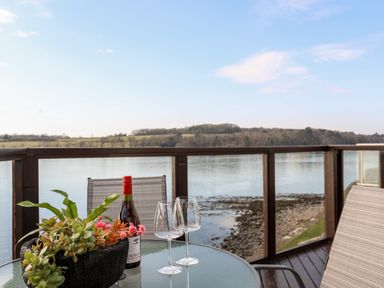 A glass table with a bottle of wine two wine glasses and a plant on a balcony overlooking water and hills at The Summerhouse in Y Felinheli