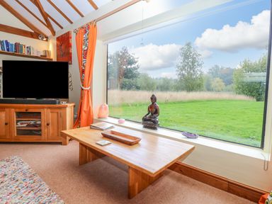A living room with a wooden coffee table a television bookshelf and large window showing a green field at The Lodge in Hickling near Catfield
