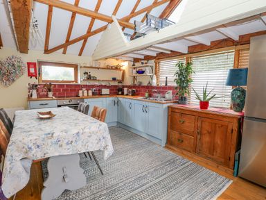 A kitchen with a dining table covered with a floral tablecloth chairs a wooden cabinet plants and a refrigerator at The Lodge Hickling near Catfield