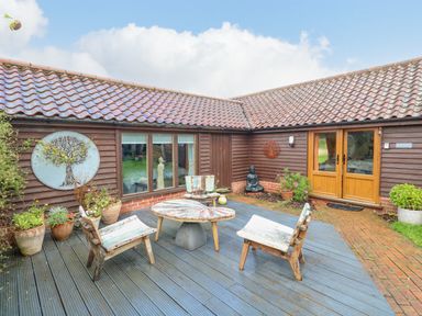 An outdoor patio with wooden chairs and a table outside brown wooden buildings at The Lodge in Hickling near Catfield