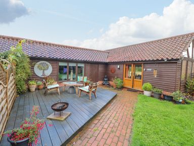 A wooden deck with chairs and a table next to a brown wooden house with a tiled roof and a brick pathway at The Lodge in Hickling near Catfield