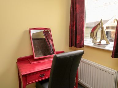 A red vanity table with a mirror and a black chair next to a window with red curtains and a silver sailboat decoration on the windowsill at Sea View Lodge in Eastfield Hall near Warkworth