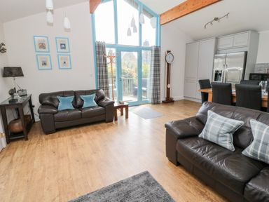 A living room with brown leather sofas and blue cushions near large blue framed windows and doors with wooden floors at Sea View Lodge in Eastfield Hall near Warkworth