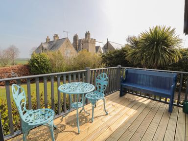 A wooden deck with a blue metal table and two chairs and a blue wooden bench with bushes and old stone buildings in the background at Sea View Lodge Eastfield Hall near Warkworth