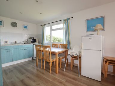 A kitchen with blue cabinets a dining table with chairs a white refrigerator and a window with curtains at Trevone - Chalet 130 in St Merryn near Padstow