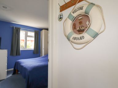 A bedroom with a blue bedspread a window with curtains a white chest of drawers and a life ring wall decoration at Trevone - Chalet 130 in St Merryn near Padstow