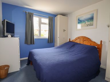 A bedroom with a wooden bed and blue bedspread next to a white wardrobe and dresser with a TV on top at Trevone - Chalet 130 St Merryn near Padstow