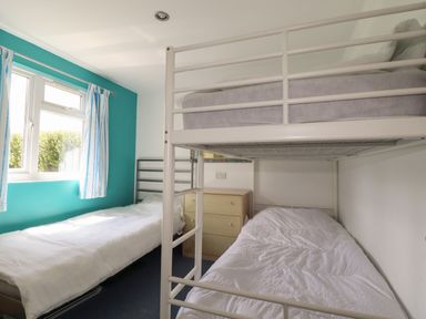 A bedroom with a bunk bed, a single bed, a window with curtains and a wooden chest of drawers at Trevone - Chalet 130 in St Merryn near Padstow