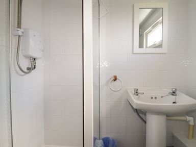 A bathroom with a shower area with an electric shower unit a pedestal sink a mirror a towel ring and a small window at Trevone - Chalet 130 St Merryn near Padstow