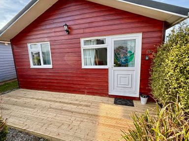A red wooden chalet with white door and windows on a wooden deck surrounded by plants at Trevone Chalet 130 St Merryn near Padstow