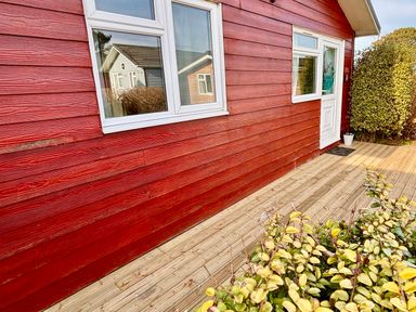 The exterior of a red wooden house with white framed windows and a white door next to a wooden deck and some bushes