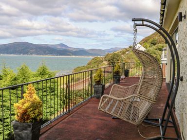A balcony with a hanging chair and potted plants overlooking the sea and hills at Machlud Haul in Llwyngwril