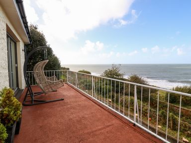 A balcony with a brown floor and hanging rattan chair overlooking the sea and green vegetation at Machlud Haul in Llwyngwril