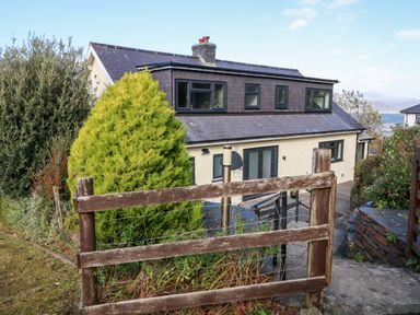 A house with dark framed windows and a wooden fence in front surrounded by greenery at Machlud Haul in Llwyngwril