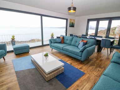 A living room with blue sofas a coffee table and large windows overlooking the sea at Machlud Haul in Llwyngwril