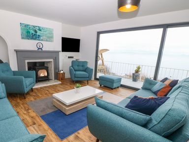 A living room with blue sofas and chairs a fireplace and a large window overlooking water at Machlud Haul in Llwyngwril