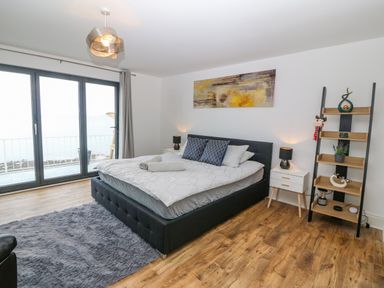 A bedroom with a large bed bedside tables lamps a standing shelf and large windows overlooking water at Machlud Haul in Llwyngwril