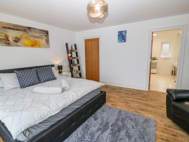 A bedroom with a large bed two pillows bedside table and lamp wooden door open door to bathroom and black armchair at Machlud Haul in Llwyngwril