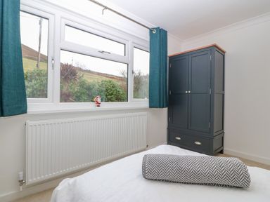 A bedroom with a window with blue curtains a gray wardrobe and a bed with a rolled blanket at Machlud Haul in Llwyngwril