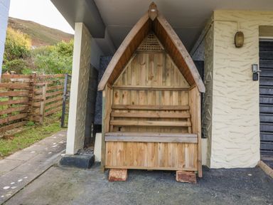 A wooden shelter bench outside a building with a textured wall next to a wooden fence at Machlud Haul in Llwyngwril