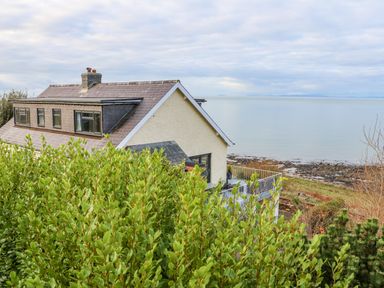 A house with a tiled roof overlooking a rocky shoreline and calm sea at Machlud Haul in Llwyngwril