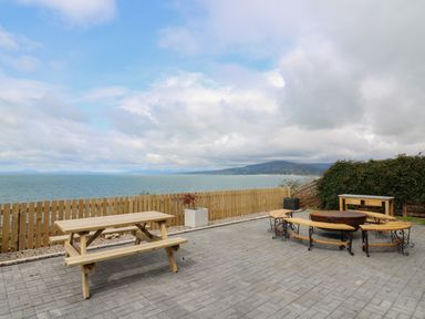 An outdoor patio with wooden picnic table fire pit with curved benches wooden table and a wooden fence by the sea at Machlud Haul in Llwyngwril