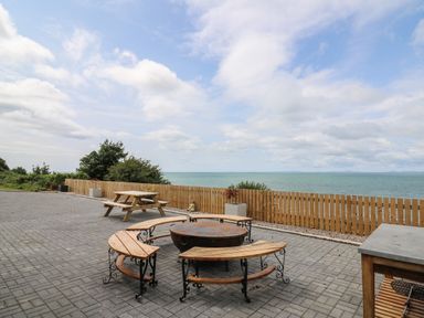 An outdoor patio area with a round fire pit surrounded by curved wooden benches a wooden picnic table a wooden fence and the sea in the background at Machlud Haul in Llwyngwril