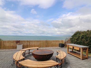An outdoor patio with a round fire pit surrounded by curved wooden benches next to a wooden table overlooking the sea at Machlud Haul in Llwyngwril