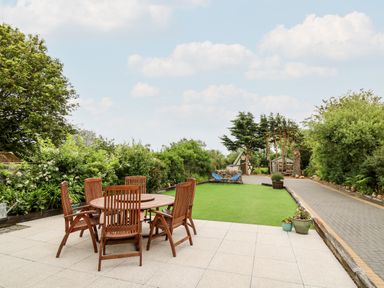 An outdoor patio with a round wooden table and six chairs next to a lawn and a paved pathway at Dolwern in Dinas Cross