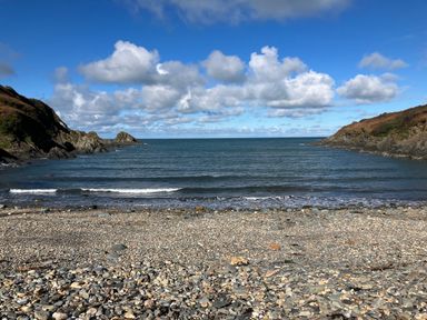 A pebble beach with calm sea between two rocky cliffs under a partly cloudy sky at Dolwern in Dinas Cross