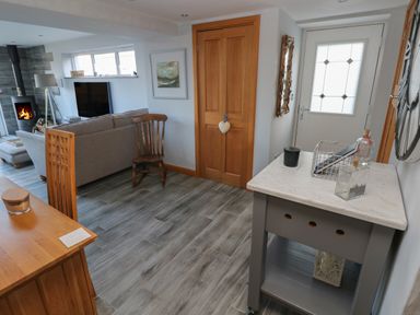 An entryway with a wooden door a chair a grey cart with bottles and a basket and a living room with a sofa and a wood stove at Dolwern in Dinas Cross