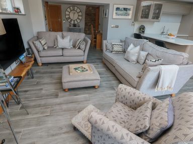 A living room with grey sofas a wooden table television and kitchen area at Dolwern in Dinas Cross