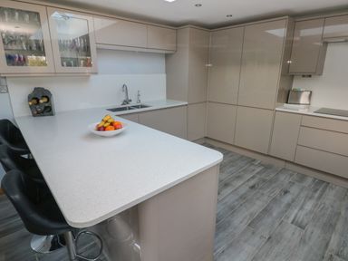 A kitchen with beige cabinets a white countertop a bowl of fruit three black bar stools and a wine rack at Dolwern in Dinas Cross