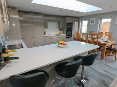 A kitchen with a white countertop island and black bar stools next to a dining table with chairs at Dolwern in Dinas Cross