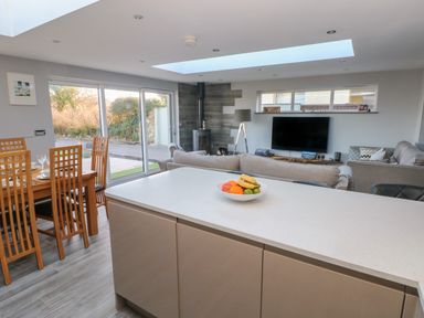 A kitchen island with fruit bowl and an open living room with sofas dining table and sliding glass doors at Dolwern in Dinas Cross