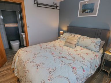 A bedroom with a bed floral bedding side tables with lamps a framed picture and an open door to a bathroom at Dolwern in Dinas Cross