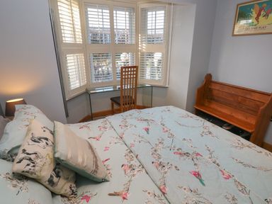 A bedroom with a bed and patterned bedding a wooden bench a glass desk with a wooden chair and windows with white shutters at Dolwern in Dinas Cross