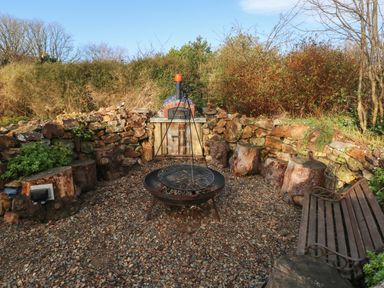 An outdoor area with a fire pit grill log seats a stone wall a wooden bench and a pizza oven at Dolwern in Dinas Cross