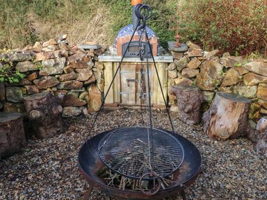 An outdoor fire pit with a hanging grill surrounded by wooden stump seats and a stone wall patio at Dolwern in Dinas Cross