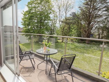 A balcony with a small round table and two chairs overlooking a grassy garden with trees at 17 Coedrath Park in Saundersfoot
