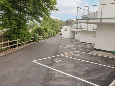 An outdoor parking lot with numbered spaces next to a white building with glass balconies and trees along a wooden fence at 17 Coedrath Park in Saundersfoot