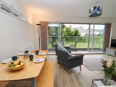 A dining table set with cups plates and fruit bowl beside a sitting area with two armchairs and a balcony with outdoor chairs and a table at 17 Coedrath Park in Saundersfoot