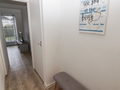 A hallway with wooden flooring a gray bench and a wall sign next to a doorway showing a room with a television and vertical blinds at 28 Coedrath Park in Saundersfoot
